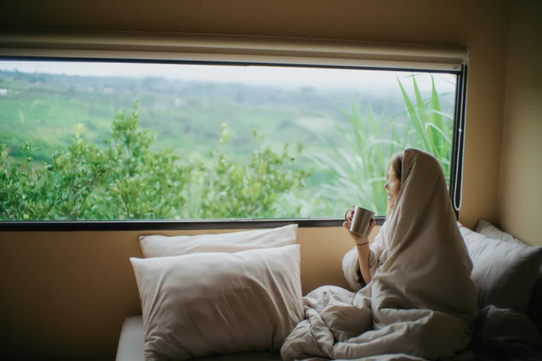 A guest wrapped in a blanket, enjoying a morning coffee while gazing out the window at the beautiful natural scenery.