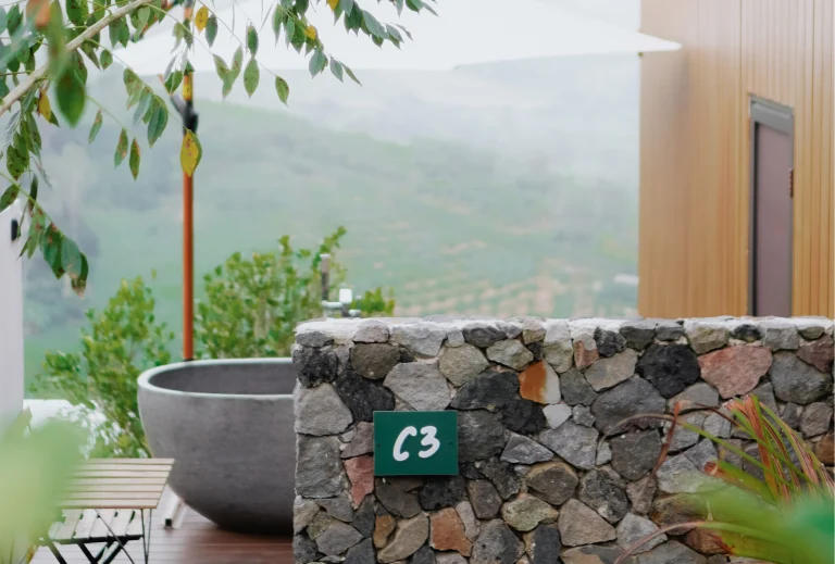 A tranquil outdoor setting with a stone bathtub surrounded by lush greenery and a view of the mountains. The bath is covered with a parasol for shade, creating the perfect relaxation spot.