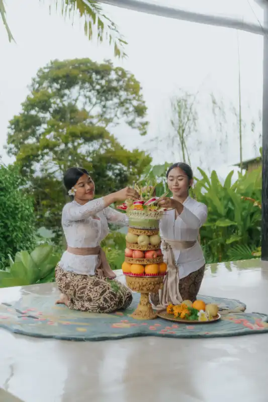 Two Balinese women in traditional kebaya attire kneel on a mat, working together to prepare a tall 'Gebogan' fruit offering for the Galungan ceremony.